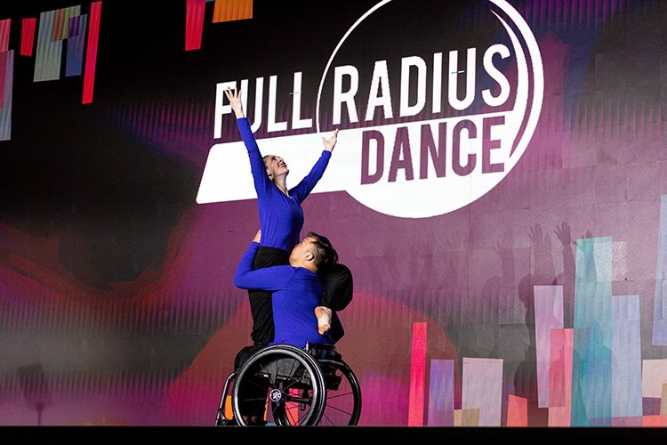 A dancer with her arms outstretched by held by another dancer in a wheelchair
