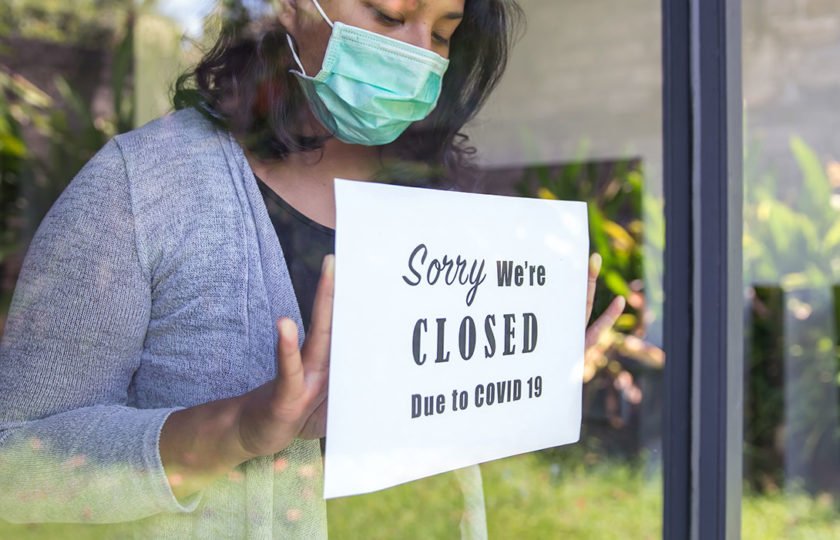 Woman with a mask on holding up a sign that says "Sorry we're closed due to COVID-19"