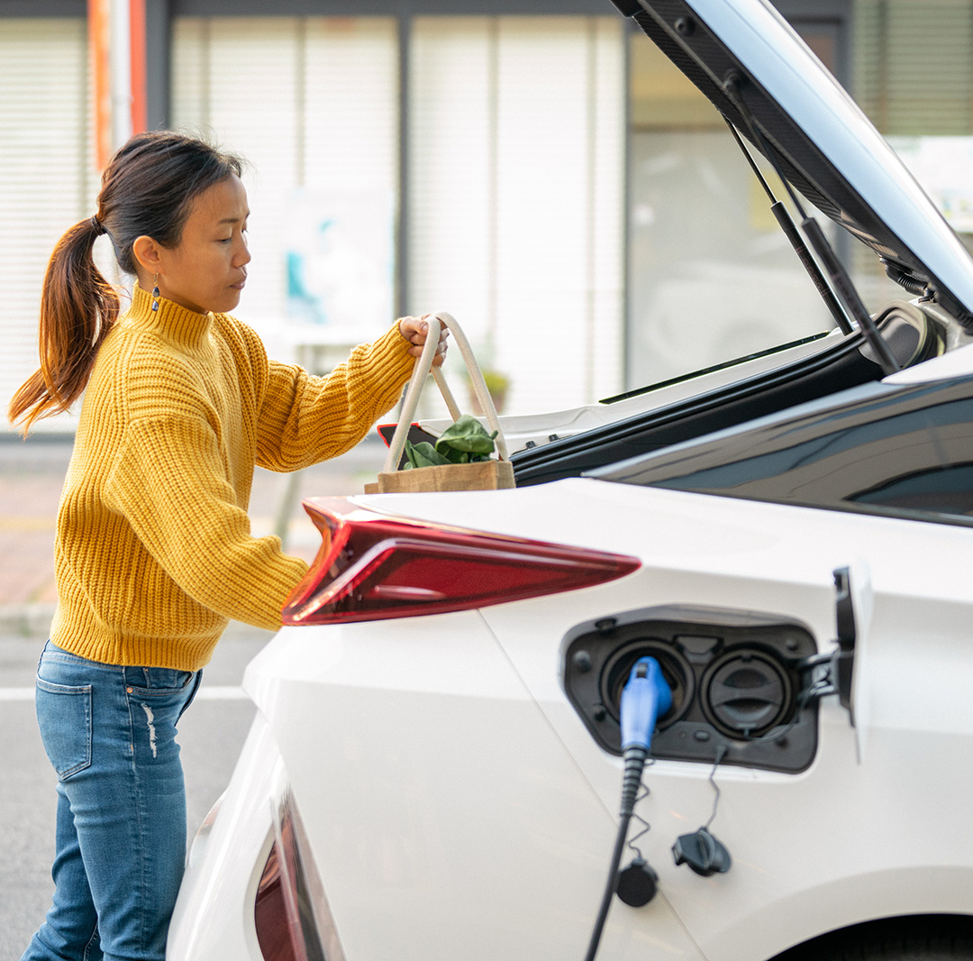 Young woman placing groceries in the back hatch of an electric vehicle while it is charging.